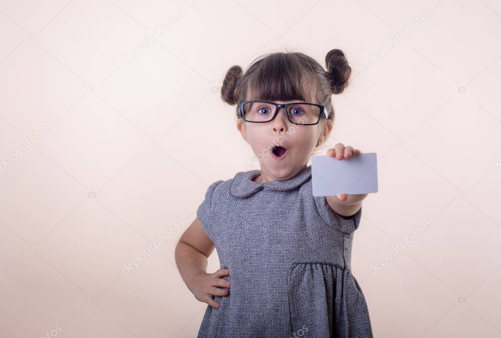 Wow! Happy shocked child holding discount white card in her hands. Surprised kid with credit card. Little girl with glasses showing empty blank paper note copy space.