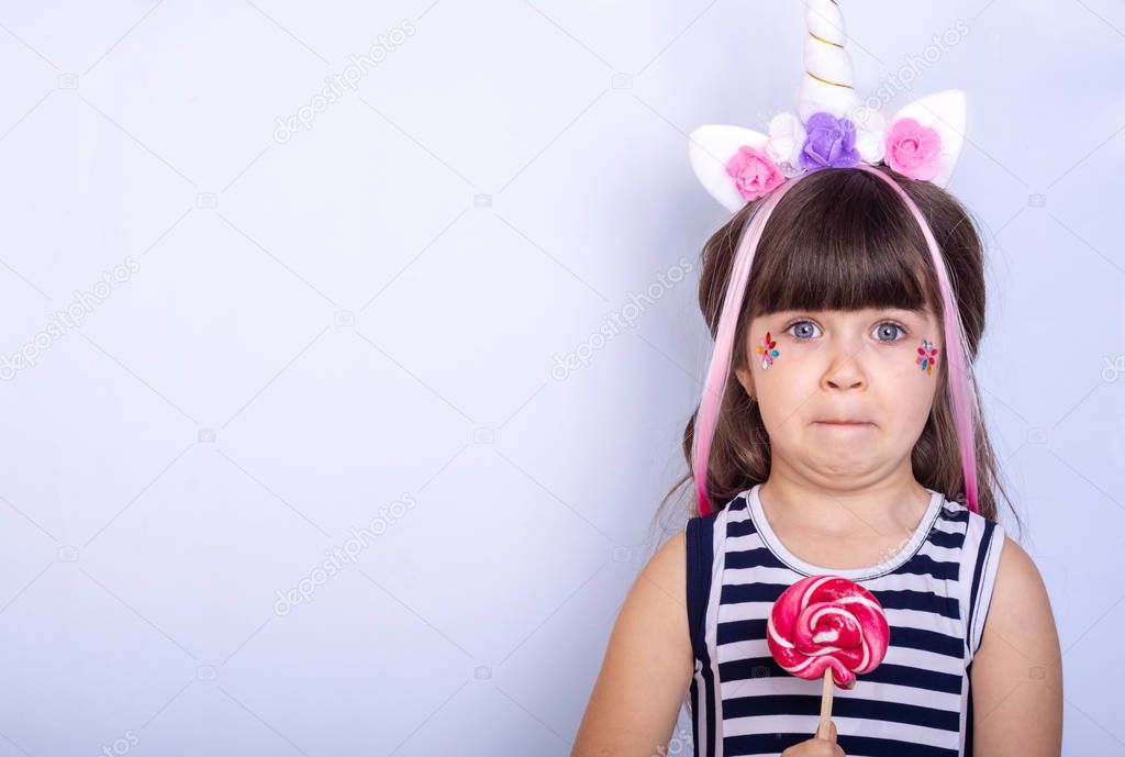 Background with funny delight child. Little girl in hair hoop with unicorn horn a holding candy. Children on empty blue banner. The concept advertising of children's goods. 