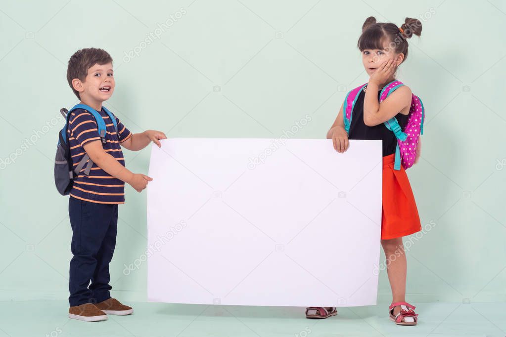 Back to school - advertisements. School Kids with backpacks holding white blank or white card. Happy children ready for school isolated on green background. 