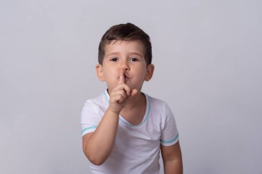 Gürültüye neden olmasın, shh. Sevimli çocuğun işaret parmağını ağzın üzerinde tutması, sus ve olumlu ifade deyip gri duvarın üzerinde durması portresi
