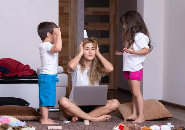Young mom in shock working on computer with unhappy kids. Exhausted mother