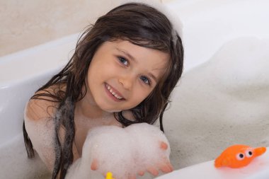 Çocuk köpük banyosu yapıyor. Çocuklar küvette banyo yapıyor. Küçük kız suyla oynuyor. Köpük banyosunda plastik ördek 