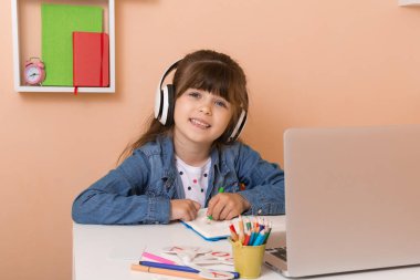 Evde sanal kurslarla çocuk eğitimi. Birincil online sınıf için etkileşimli içerik. Evde öğrenim. Masada dizüstü bilgisayar kullanan kulaklıklı kız.