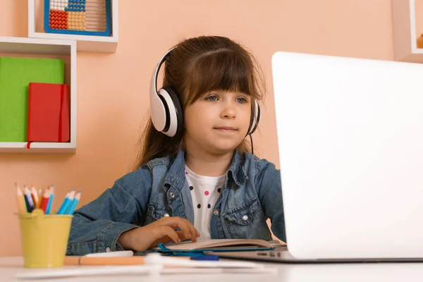 Evde sanal kurslarla çocuk eğitimi. Birincil online sınıf için etkileşimli içerik. Evde öğrenim. Masada dizüstü bilgisayar kullanan kulaklıklı kız.