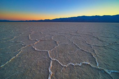 Çatlak tuz oluşumları Badwater Havzası boyunca uzanır Ölüm Vadisi Nevada 'da altın saat, uzak dağlar siluetli ve canlı bir günbatımı gökyüzü sakin ve dramatik bir çöl manzarası yaratır..