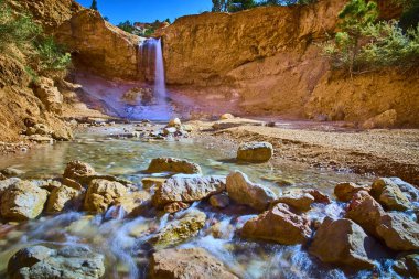 Bryce Canyon Ulusal Parkı 'ndaki yosunlu mağara patikası boyunca sakin bir manzarada Utah, Tropic Ditch Falls' u kızıl uçurumların üzerinden seyrek çöl bitkileriyle çevrili kayalık bir dereye döküyor..