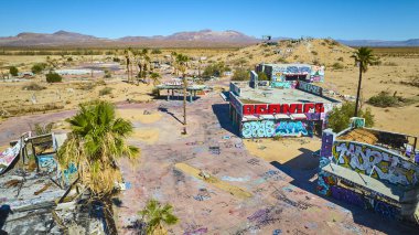 Kaliforniya çölündeki terk edilmiş bir su parkının havadan görünüşü. Çürüyen yapılar ve palmiye ağaçları üzerinde canlı grafitiler var. Açık mavi gökyüzü altında geniş kurak bir araziyle çevrili..