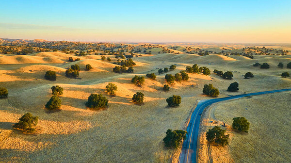 Вид с воздуха на Хот Спрингс Роуд, извивающийся через золотые холмы и дубы в Калифорнии в течение золотого часа с яркими цветами заката заполняя мирный сельский пейзаж.