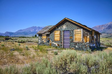 Terk edilmiş bir ev Lee Vining California yakınlarındaki kuru tarlaların arasında duruyor. Williams Butte ve engebeli dağlar açık mavi gökyüzünün altında yükseliyor..