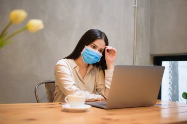 Maskeli güzel bir kadın kahve dükkanında bilgisayarla çalışıyor. 