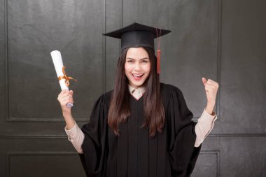 Mezuniyet elbisesi içinde siyah arka planda gülümseyen ve tezahürat yapan genç bir kadının portresi. 