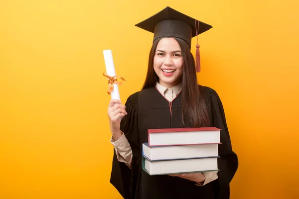 穿着毕业礼服的漂亮女人拿着黄色背景的书和证书 — 图库照片