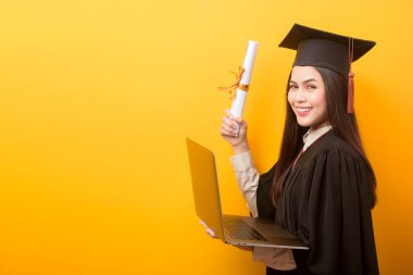 Mezuniyet elbisesi giymiş güzel bir kadının portresi sarı arka planda dizüstü bilgisayarı tutuyor. 