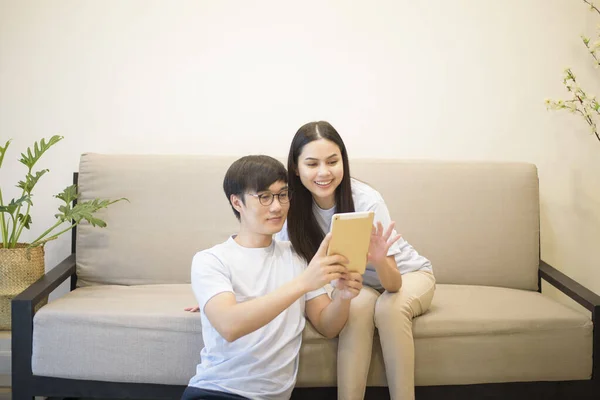 a happy couple wearing blue shirt is relaxing and holding a tablet on a sofa at home