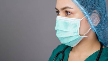 Yeşil önlüklü genç, kendine güvenen bir kadın doktor gri arka plan stüdyosunun üzerinde cerrahi maske takıyor.