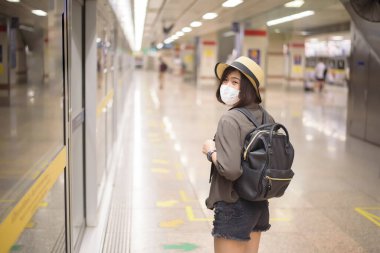 Genç ve güzel bir Asyalı kadın koruyucu maske takıyor metro istasyonunda duruyor, yeni normal seyahat, covid-19 koruma, güvenli seyahat, COVID-19, salgın sonrası seyahat konsepti.