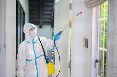 PPE kostümlü bir tıbbi personel oturma odasında dezenfektan spreyi kullanıyor, Covid-19 koruma, dezenfeksiyon konsepti. .