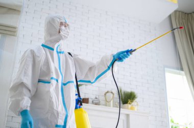 PPE kostümlü bir tıbbi personel oturma odasında dezenfektan spreyi kullanıyor, Covid-19 koruma, dezenfeksiyon konsepti. .