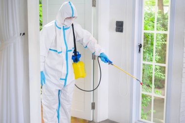 PPE kostümlü bir tıbbi personel oturma odasında dezenfektan spreyi kullanıyor, Covid-19 koruma, dezenfeksiyon konsepti. .