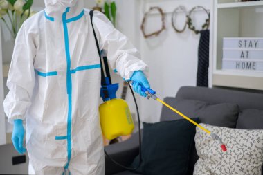 PPE kostümlü bir tıbbi personel oturma odasında dezenfektan spreyi kullanıyor, Covid-19 koruma, dezenfeksiyon konsepti. .