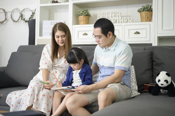 Happy asian parent teaching little cute daughter to do homework and drawing picture together at home.