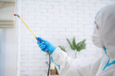 PPE kostümlü bir tıbbi personel oturma odasında dezenfektan spreyi kullanıyor, Covid-19 koruma, dezenfeksiyon konsepti. .