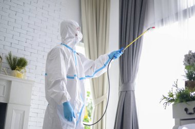 PPE kostümlü bir tıbbi personel oturma odasında dezenfektan spreyi kullanıyor, Covid-19 koruma, dezenfeksiyon konsepti. .