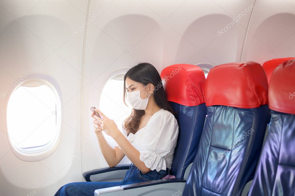 A young woman wearing face mask is using smartphone onboard, New normal travel after covid-19 pandemic concept	