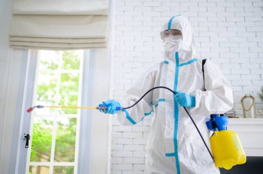 PPE kostümlü bir tıbbi personel oturma odasında dezenfektan spreyi kullanıyor, Covid-19 koruma, dezenfeksiyon konsepti. .