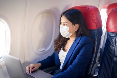 Yüz maskesi takan genç bir iş kadını uçakta dizüstü bilgisayar kullanıyor. Covid-19 salgınından sonra normal bir yolculuk.