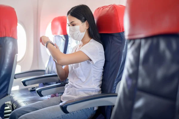 A traveling woman wearing protective mask onboard in the aircraft using smart watch, travel under Covid-19 pandemic, safety travels, social distancing protocol, New normal travel concept