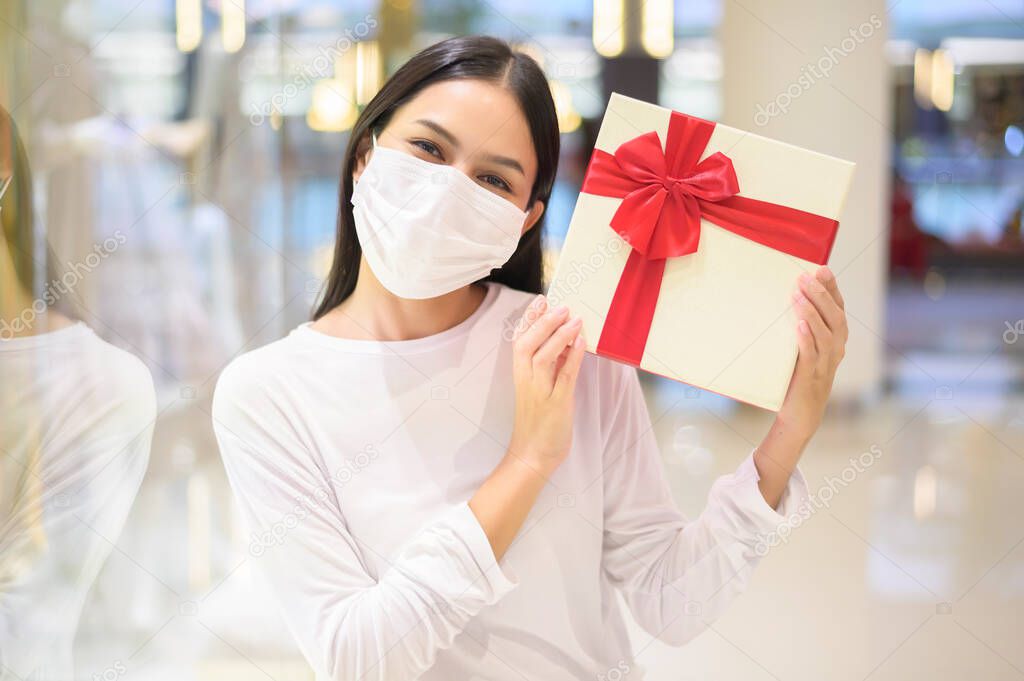 A woman wearing protective mask holding a gift box in shopping mall, shopping under Covid-19 pandemic, thanksgiving and Christmas concept.