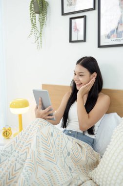 Genç Asyalı kadın yatak odasında tablet bilgisayar kullanmaktan zevk alıyor.