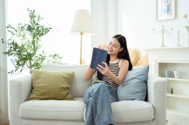 Genç Asyalı kadın evdeki oturma odasında tablet bilgisayar kullanmaktan zevk alıyor