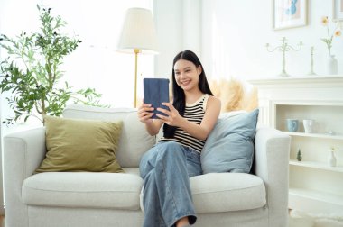 Genç Asyalı kadın evdeki oturma odasında tablet bilgisayar kullanmaktan zevk alıyor