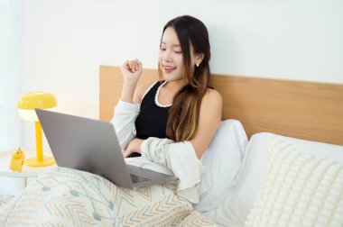 Genç Asyalı kadın yatak odasında dizüstü bilgisayar kullanmaktan zevk alıyor.