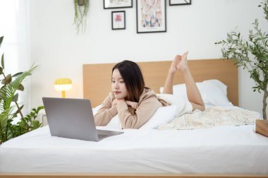 Genç Asyalı kadın yatak odasında dizüstü bilgisayar kullanmaktan zevk alıyor.