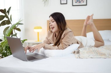 Genç Asyalı kadın yatak odasında dizüstü bilgisayar kullanmaktan zevk alıyor.