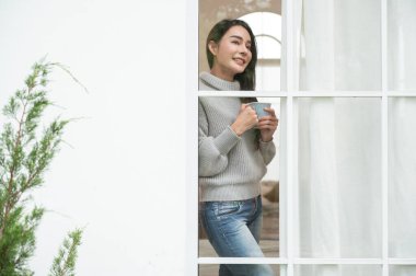 Süveter giyen bir kadın sıcak bir içecek yudumluyor, pencerenin yanında mutlu görünüyor..