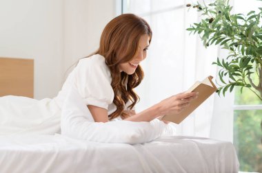 Beyaz elbiseli bir kadın, bitkilerle dolu aydınlık bir odada yatakta kitap okumaktan hoşlanır..