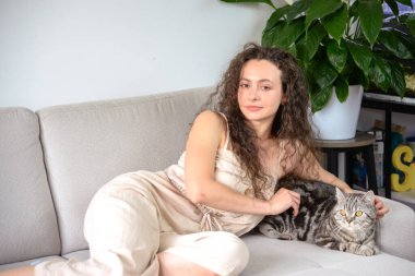 Bir kadın kedisiyle kanepede uzanıyor, yemyeşil bitkilerle çevrili, sıcak, rahat bir ortam yaratıyor.