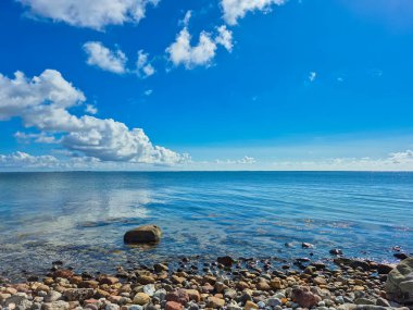 Sakin suların üzerinde berrak mavi gökyüzü ile çevrili renkli çakıl taşlarını gösteren huzurlu bir sahil manzarası.