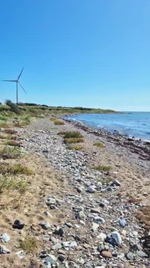 Rüzgâr türbini, kayalık kıyılar ve berrak bir gökyüzünün altında parlayan su içeren sakin bir sahil manzarası..
