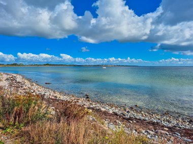 Berrak suları, kayalık kıyı şeridini ve mavi gökyüzünün altındaki canlı bulutları gösteren sakin bir sahil manzarası..