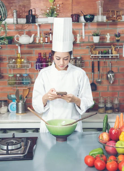 young woman chef black hair in white suit uniform look at the smartphone at kitchen room