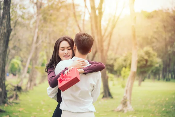 彼は公園の日没時に現在の赤いボックスの彼女を抱擁しながら美しいアジアの女性が男の肩に傾く — ストック写真