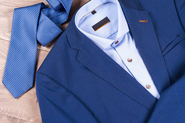 набор классической мужской одежды - синий костюм, рубашки, коричневые туфли, пояс и галстук на деревянном фоне.