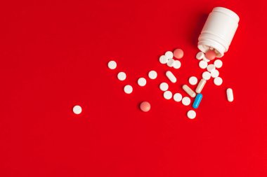 Kırmızı arka planda devrilmiş beyaz bir şişeden beyaz haplar dökülüyor.