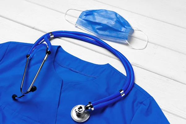 白い木製の背景に聴診器とフェイスマスクと医師の医療制服 — ストック写真