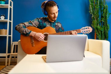 Genç beyaz adam dizüstü bilgisayarını kullanarak evde gitar dersi alıyor.